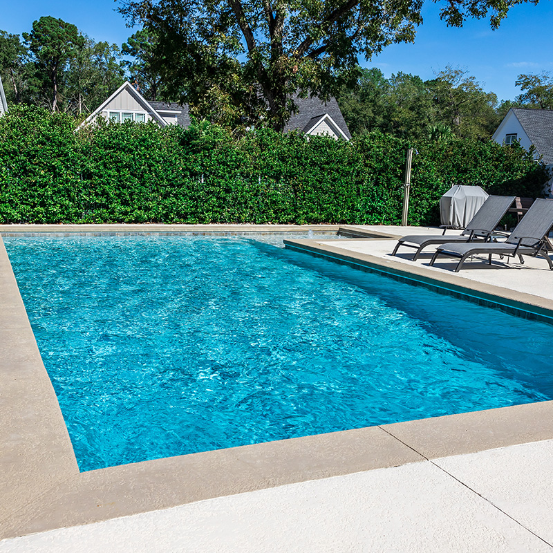 A rectangular new swimming pool with tan concrete edges in the fenced backyard of a new construction house
